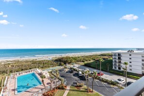 A pool with a view of the beach, a parking lot, and a building with balconies.