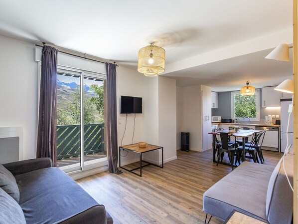 Una sala de estar moderna con sofá, una mesa de comedor y sillas. Hay una zona de cocina con microondas y pileta. Un televisor está montado en la pared y una ventana ofrece vistas al exterior.