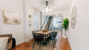 Un coin repas avec une table en bois et des chaises noires, un escalier en arrière-plan et une grande plante sur la droite.