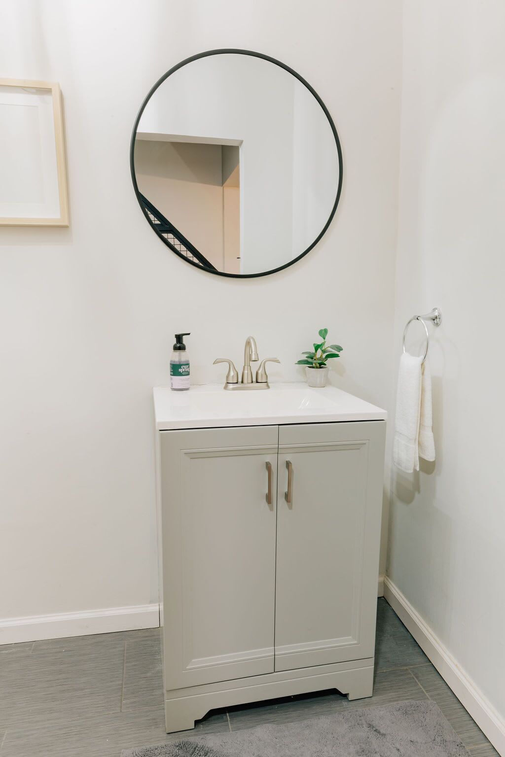 Une salle de bain avec un meuble-lavabo blanc, un miroir rond, un robinet, un distributeur de savon, une petite plante en pot et une serviette accrochée à un crochet.