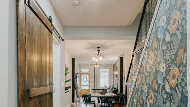 Un couloir avec un plancher en bois, un coin repas avec une table et des chaises, et un escalier dont les murs sont recouverts d’un papier peint à motifs floraux.