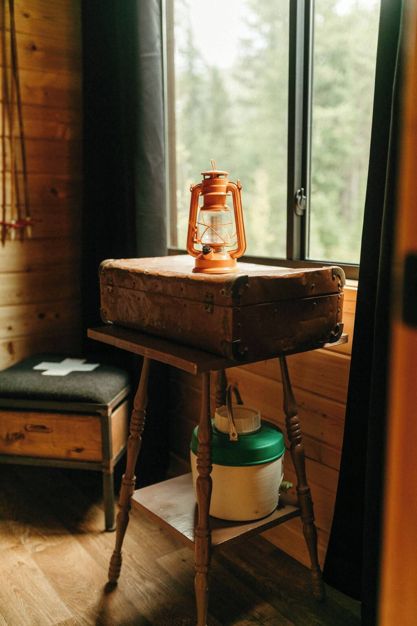 A rustic wooden cabin interior with a vintage suitcase on a wooden table, a green and white container, and a bench with a white cross.