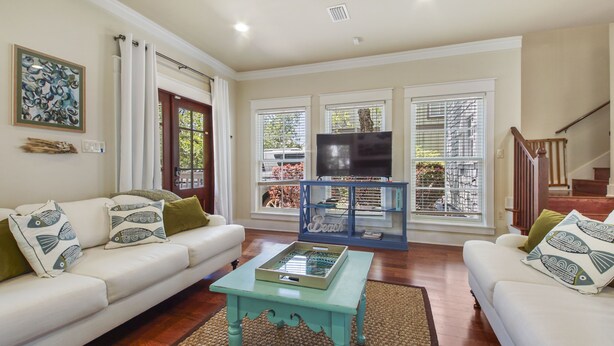 A living room with a white sofa, a coffee table, and a TV on a stand. There are large windows with blinds, a staircase, and a door with curtains. - "Will O Mist" - Inlet Beach Home, Bikes, Pool, Dock (Inlet Beach)