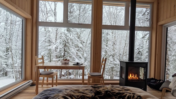 Um quarto aconchegante com uma cama, uma mesa com cadeiras, uma lareira e grandes janelas com vista para uma floresta nevada.