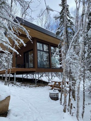 A cabin with large windows, a wooden deck, and a snow-covered landscape. - Tanières Microchalets (Baie-St-Paul)