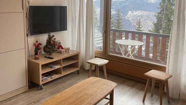 A modern living room with a wooden bench, a small table, and a TV on a wooden stand. There is a balcony with a view of mountains and a white table outside.