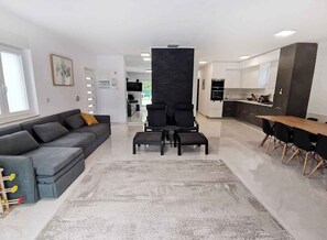 Ein modernes Wohnzimmer mit einem grauen Sofa, schwarzen Sesseln und einem Couchtisch. Es gibt eine Küchenzeile mit einem Esstisch und Stühlen.