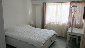 A single-bed room with a white bed, a small bedside table with a lamp, a desk with a chair, and a window with sheer curtains.