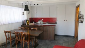 A compact kitchen with a dining area, featuring a wooden table and chairs, a red backsplash, and a hanging pendant light.