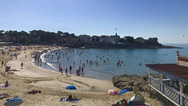 A beach with people swimming and sunbathing, a pier with umbrellas, and buildings in the background.
