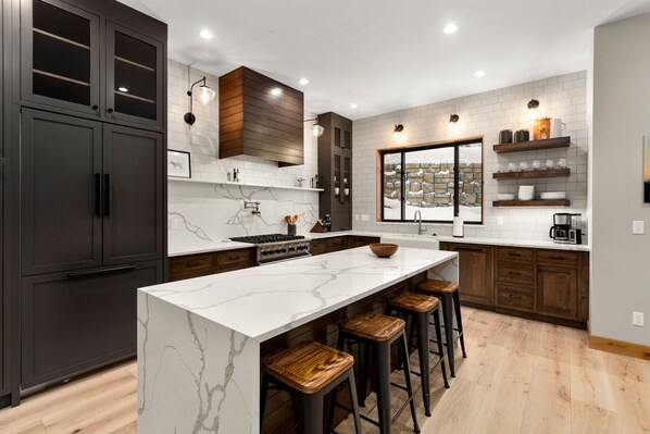 A modern kitchen with dark cabinets, a marble island, wooden stools, and a stone backsplash.
