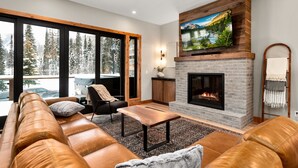 A modern living room with a fireplace, leather sofa, and a large window overlooking a snowy landscape. - Mountain modern, brand new luxury ski in and ski out townhome on Whitefish Mountain Resort! (Whitefish)
