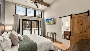 A spacious bedroom with a large bed, a ceiling fan, a sliding barn-style door, and a view of the outdoors through a large window. - Mountain modern, brand new luxury ski in and ski out townhome on Whitefish Mountain Resort! (Whitefish)