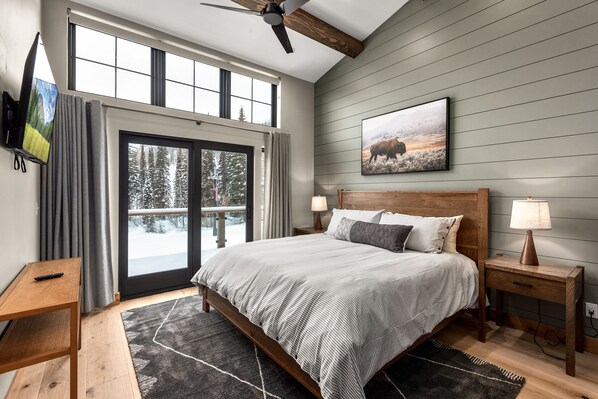 A bedroom with a large bed, a wooden headboard, a nightstand with a lamp, a TV mounted on the wall, and a window with curtains.