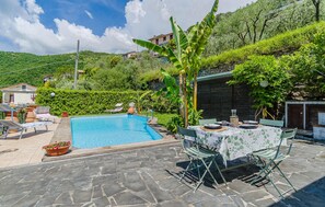 A swimming pool with lounge chairs and a table set up for dining, surrounded by lush greenery and a stone patio. - Gorgeous home in Testana (Testana)