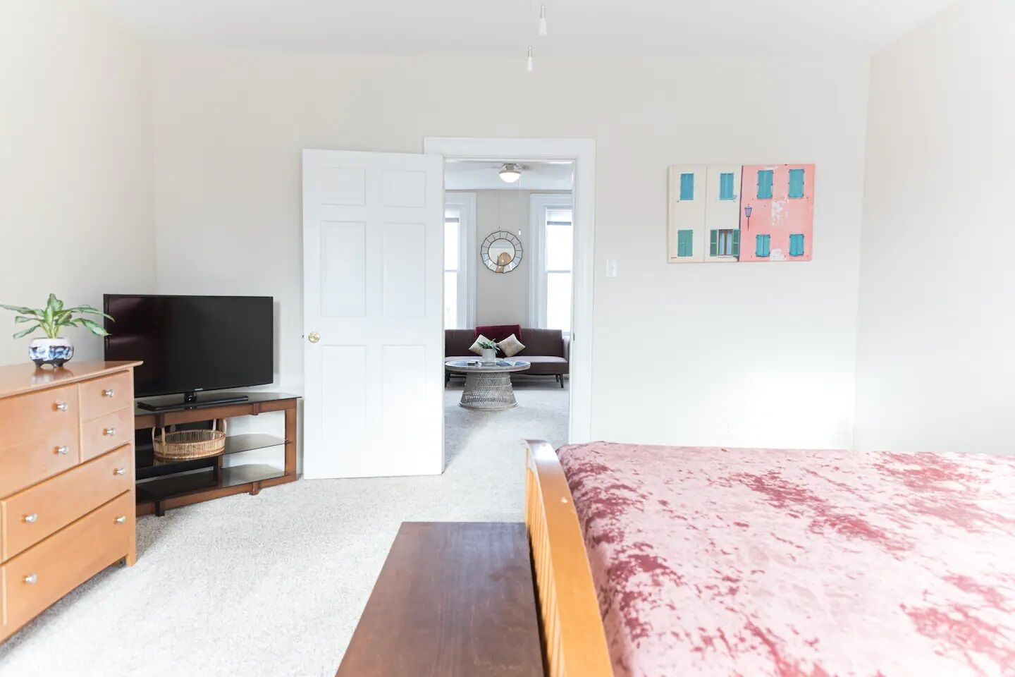 A bedroom with a bed, a TV, a dresser, and a door leading to a living area with a sofa and a coffee table.