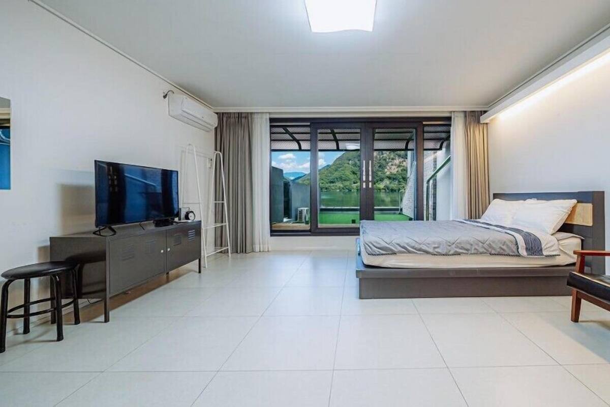 A modern hotel room with a bed, a TV, a desk, and a view of a lake.