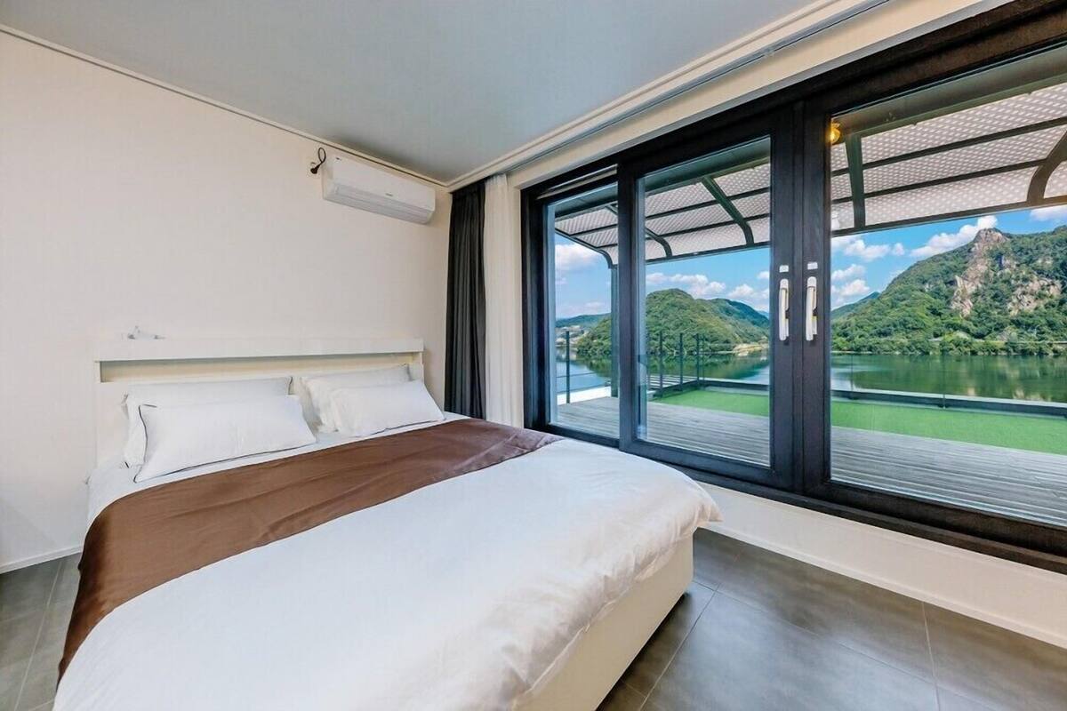 A modern hotel room with a large bed, a view of a lake and mountains through floor-to-ceiling windows, and a built-in air conditioning unit.