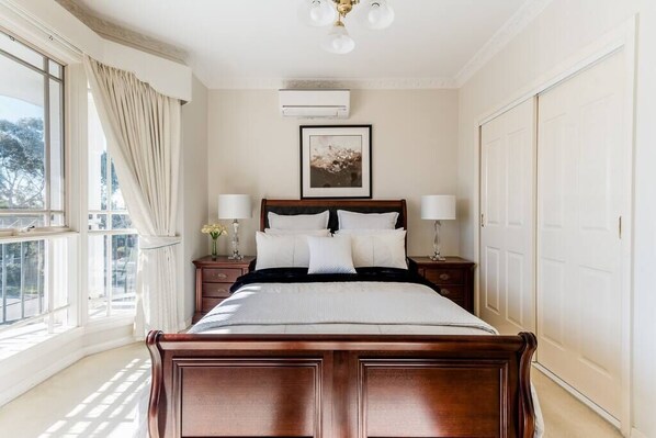 A bedroom with a large bed, two bedside tables, a window with curtains, an air conditioning unit, and a framed picture on the wall.