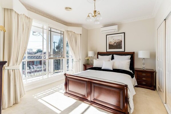 Un dormitorio amplio con una cama grande, una mesita de noche, un buró, una ventana con cortinas y una araña.