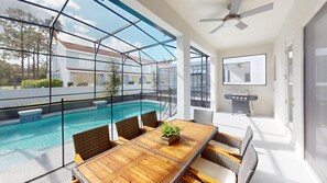 A modern outdoor pool area with a wooden table and wicker chairs, a barbecue grill, and a ceiling fan. - Bernie Pool Home 8 BED (Orlando)