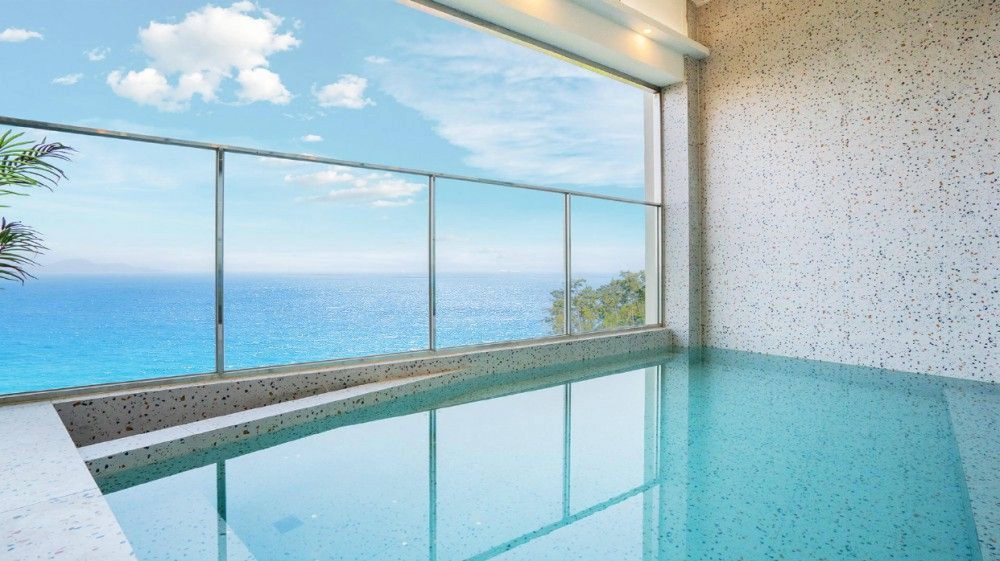 A clear view of a swimming pool with a glass barrier, a view of the ocean, and a speckled wall.
