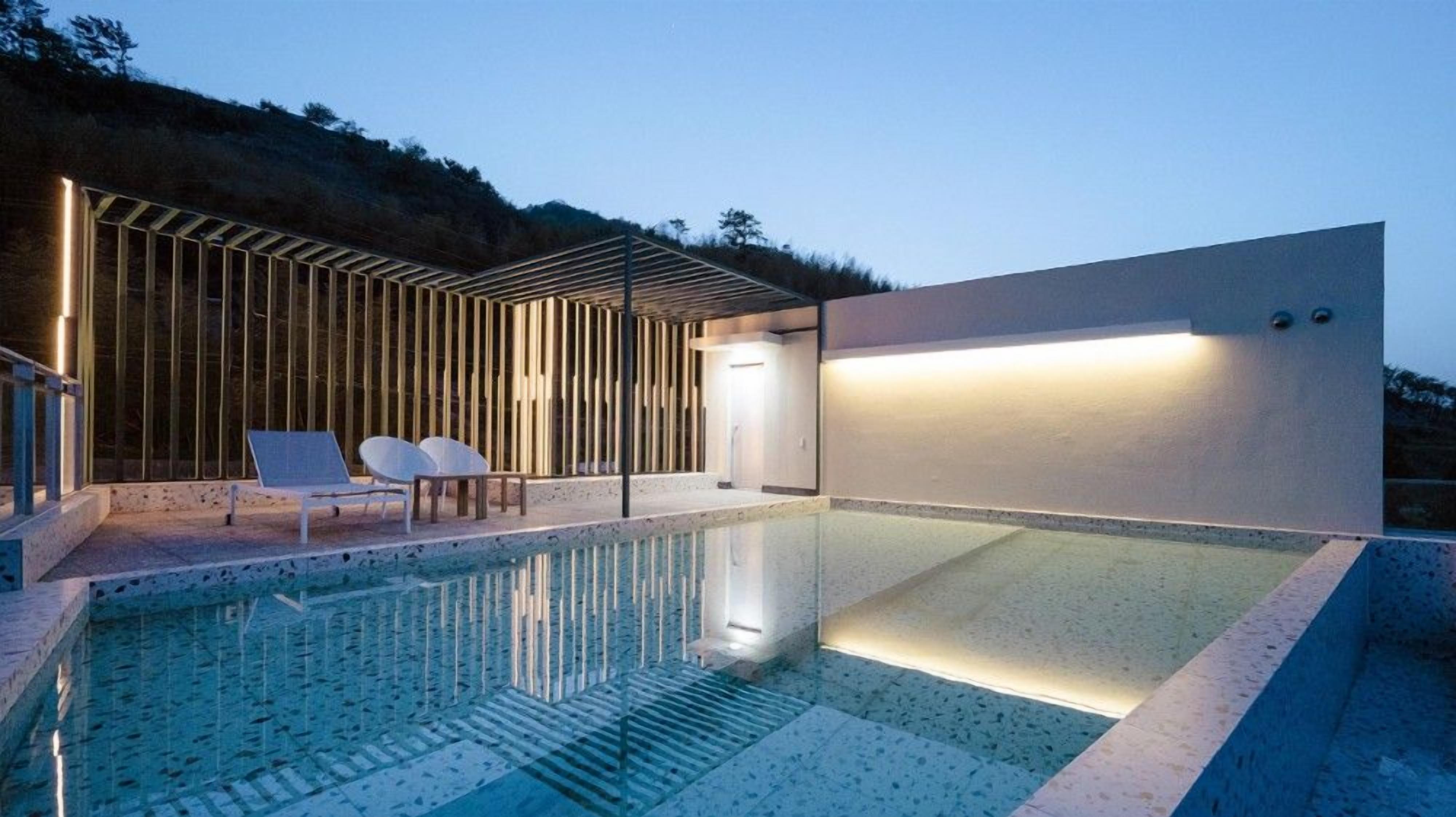 A modern outdoor pool area with a clear glass surface, a wooden pergola, and two white lounge chairs.