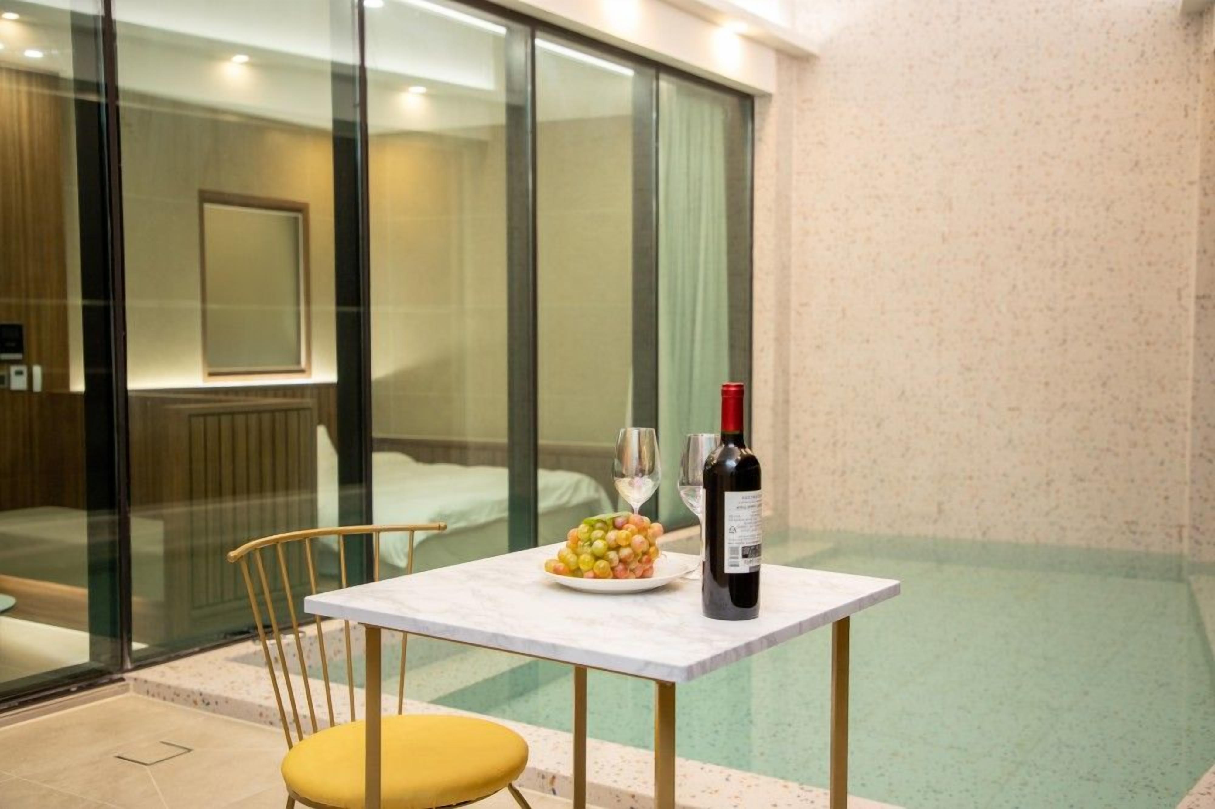 A hotel room with a glass partition, a bed, a small table with a bottle of wine and a plate of fruit, and a yellow chair.
