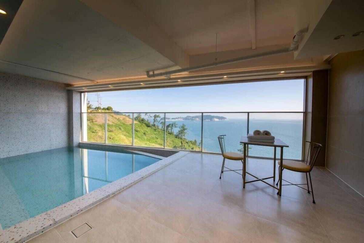 A modern indoor swimming pool with a view of the sea and hills.