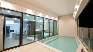 A modern indoor swimming pool with glass walls, a kitchen area visible through the glass, and a spa room with a bed.