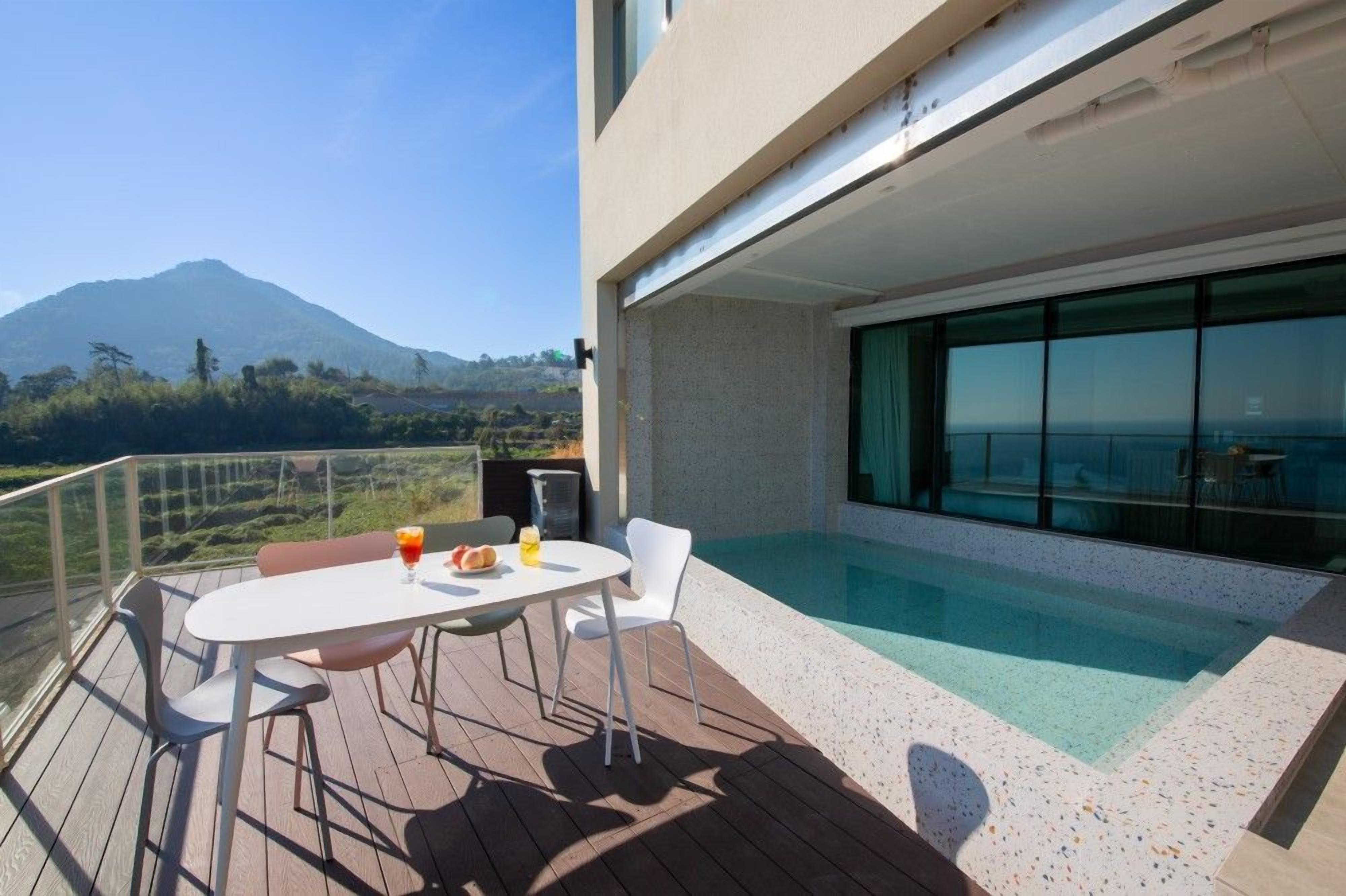 Un área de piscina moderna al aire libre con una mesa y sillas, barandilla de cristal y vista a la montaña.