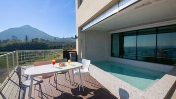Un área de piscina moderna al aire libre con una mesa y sillas, barandilla de cristal y vista a la montaña.