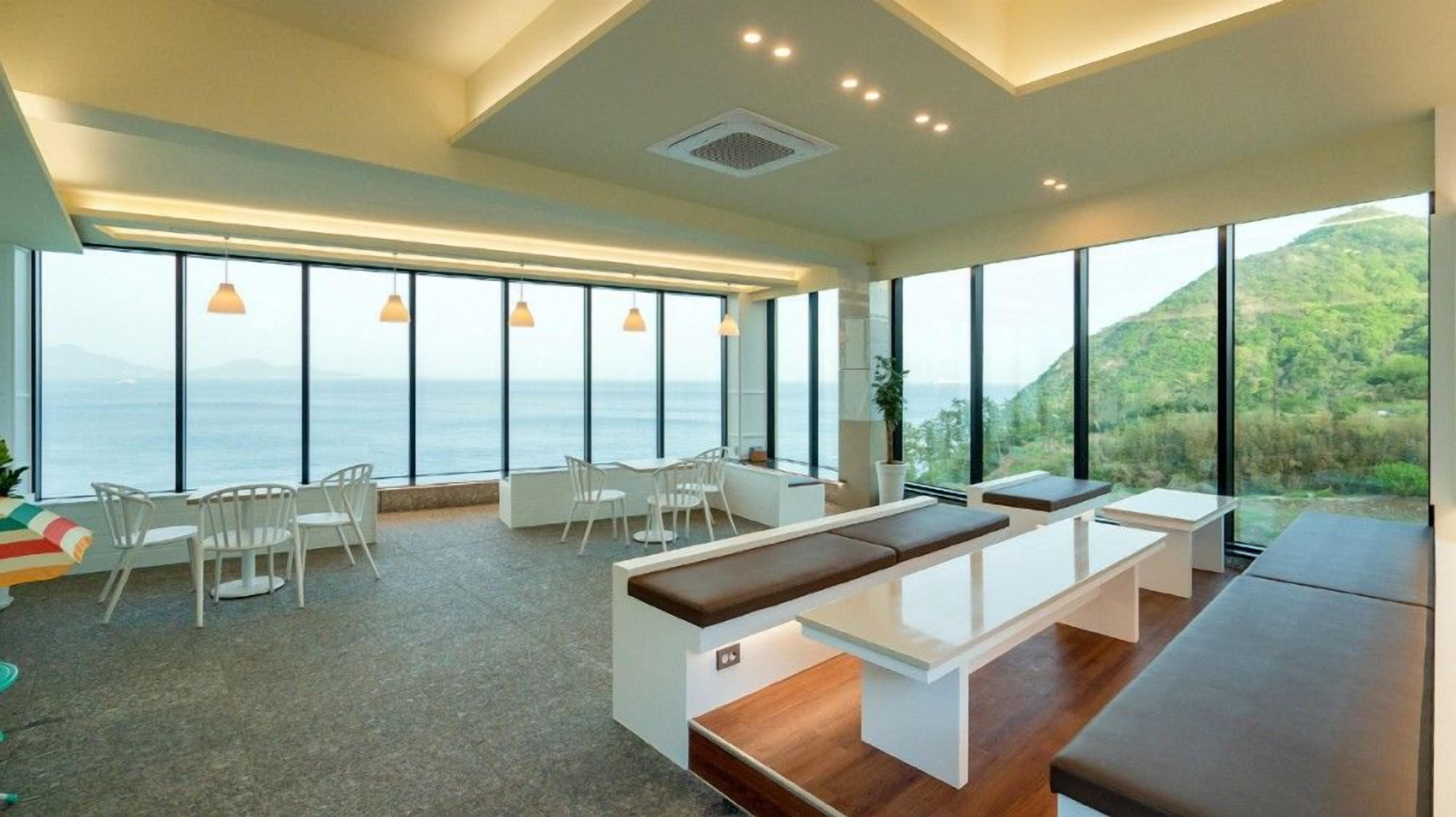Un interior moderno con ventanas amplias que ofrecen vistas al mar y a las colinas. Hay sillas blancas y un largo banco.