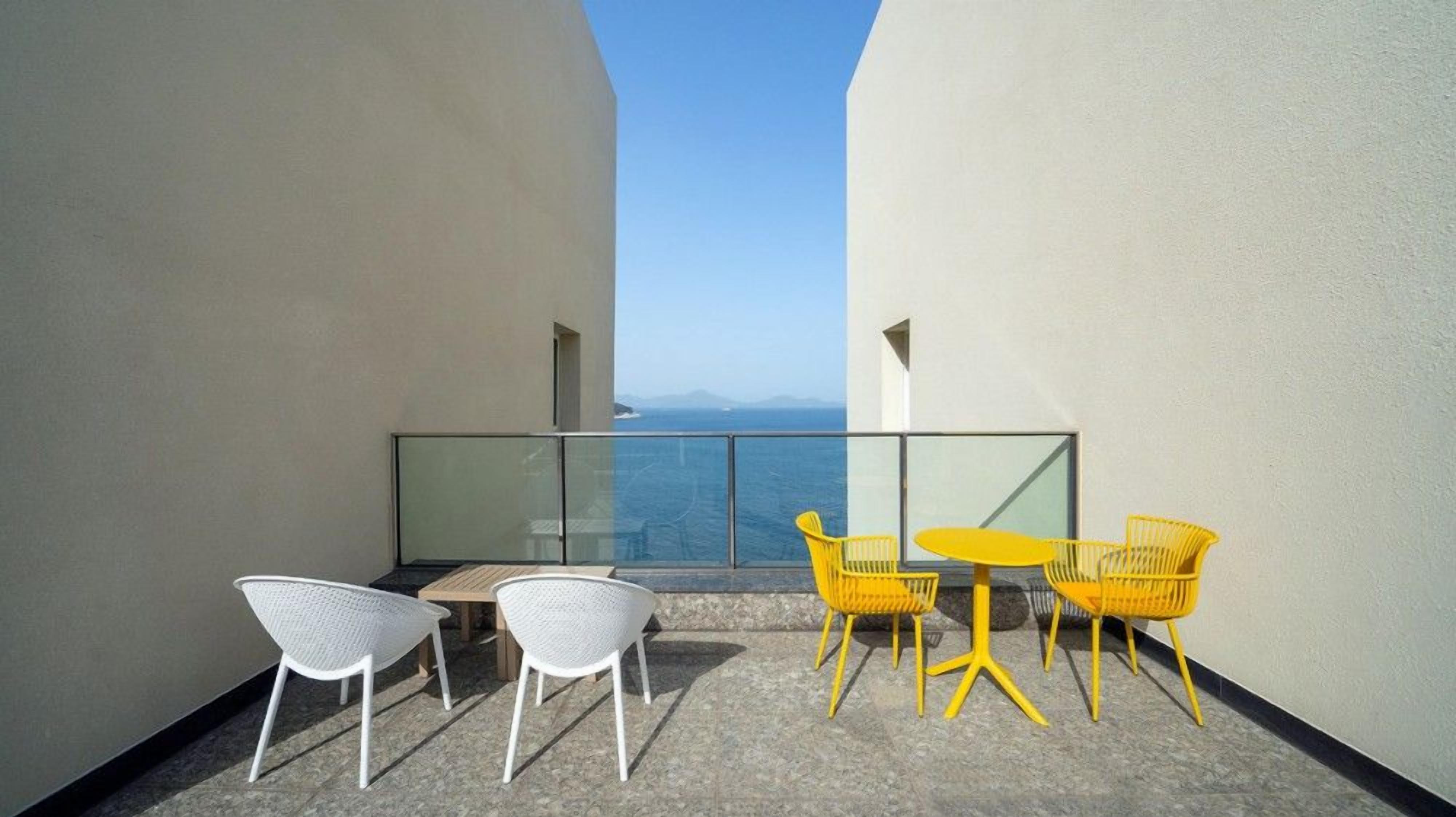 Un balcone con sedie bianche e gialle, un tavolino e una ringhiera in vetro con vista sul mare.