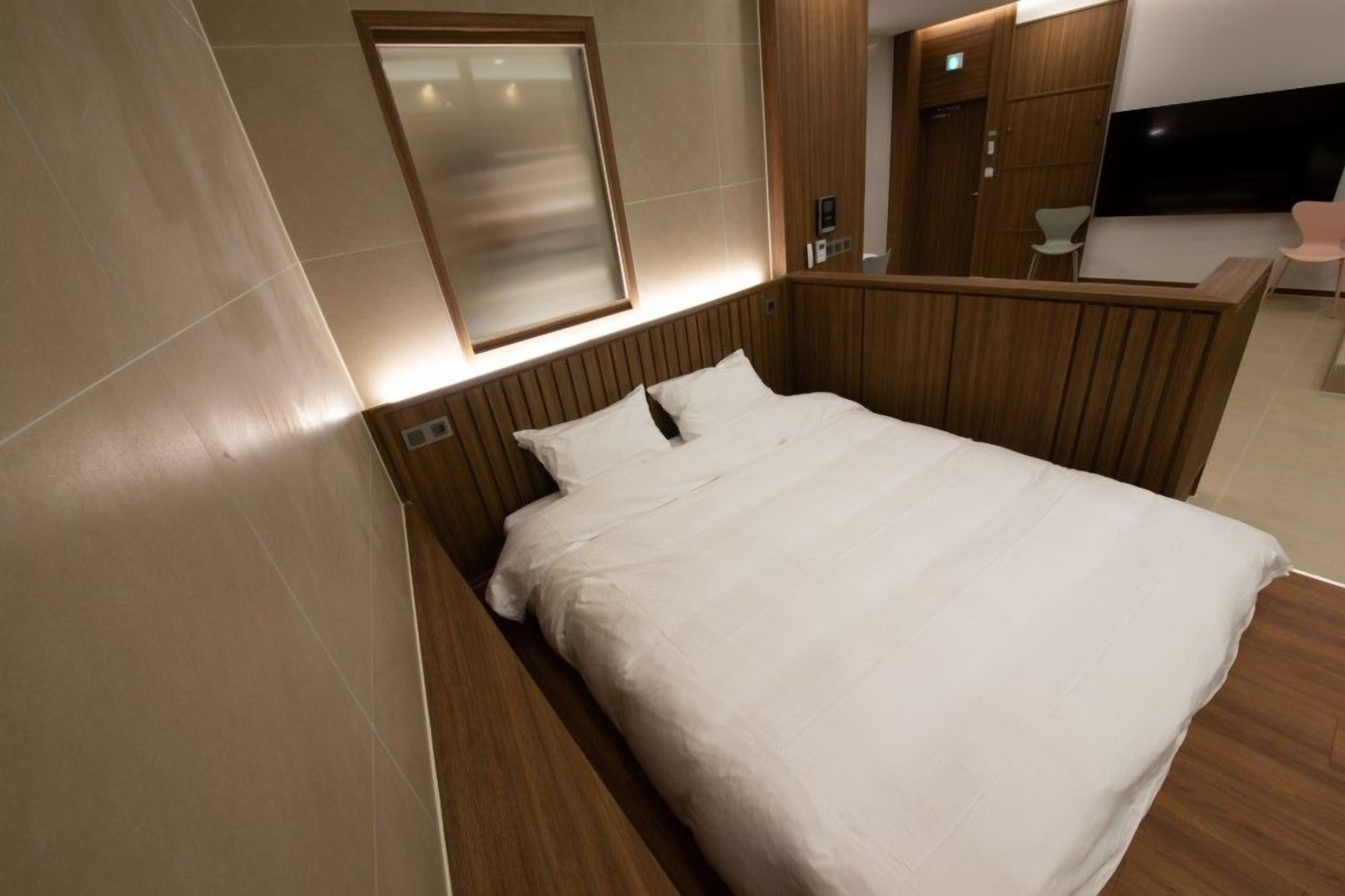 A modern hotel room with a wooden headboard, a large bed with white linens, a wall-mounted TV, and a small seating area with a chair.