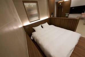 A modern hotel room with a wooden headboard, a large bed with white linens, a wall-mounted TV, and a small seating area with a chair. - The Beige (Geoje)