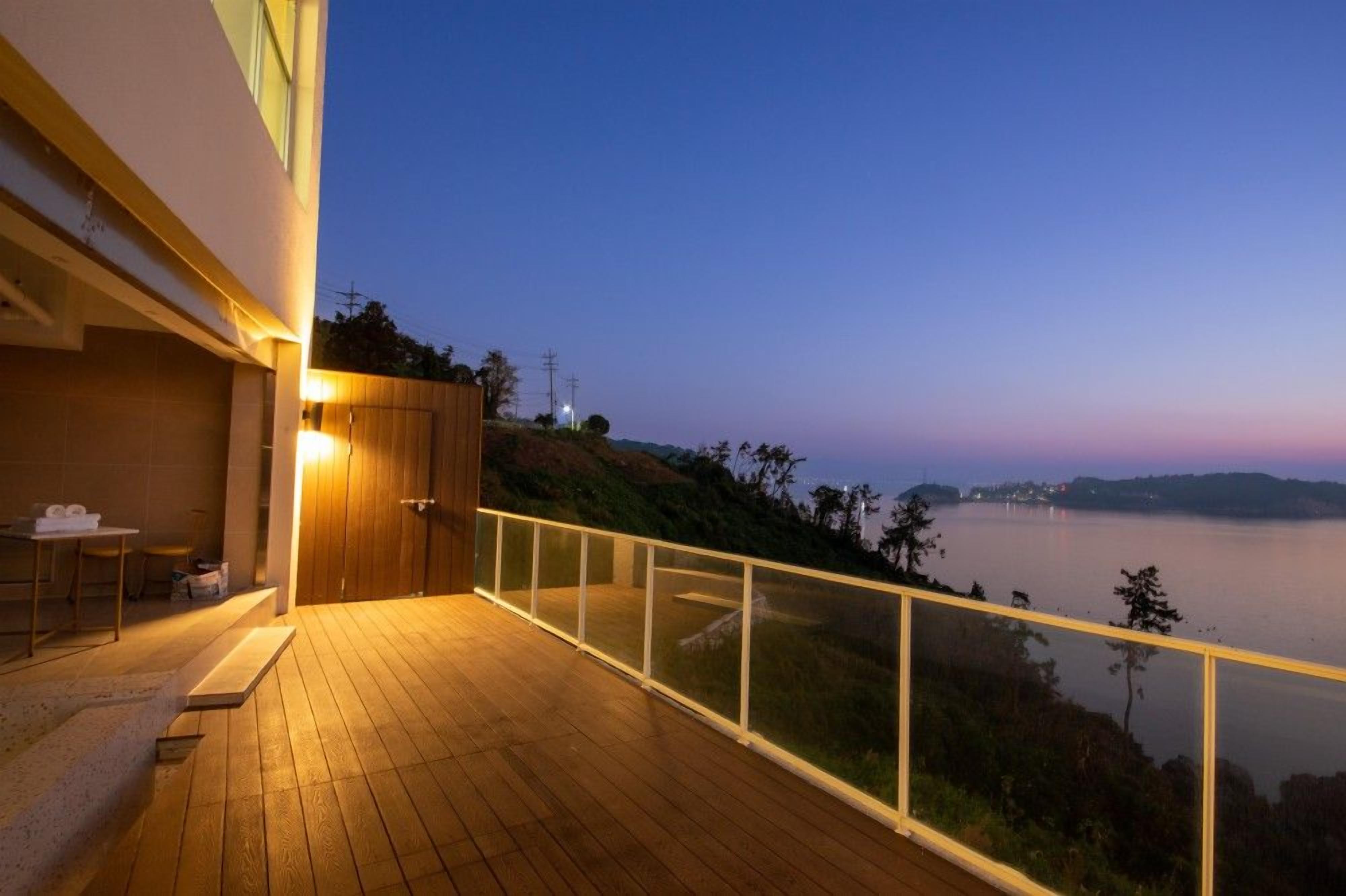 Eine Holzterrasse mit Glasgeländer, die auf ein Gewässer und Hügel blickt.