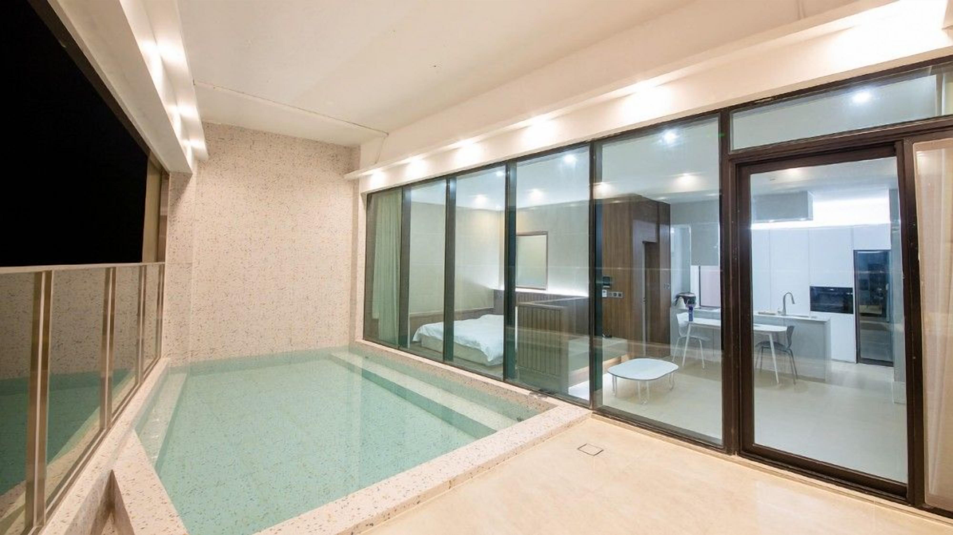 Quarto de hotel moderno com varanda envidraçada, uma pequena piscina e cozinha americana visíveis através das portas de vidro.