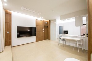 A modern kitchen with a built-in oven, a dining area with a white table and chairs, and a flat-screen TV mounted on the wall. - The Beige (Geoje)