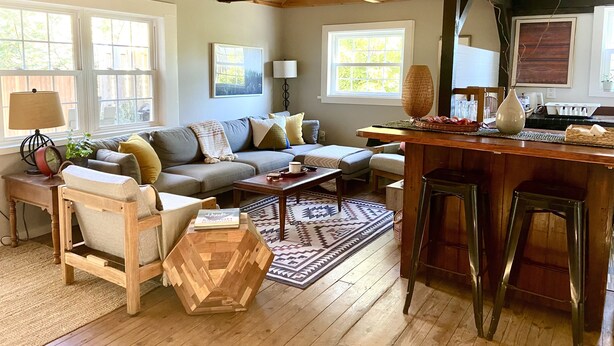 A cozy living room with a sofa, coffee table, and wooden accents. There are windows, a lamp, and a rug. - Modern Rustic Retreat in Poconos (Greentown)