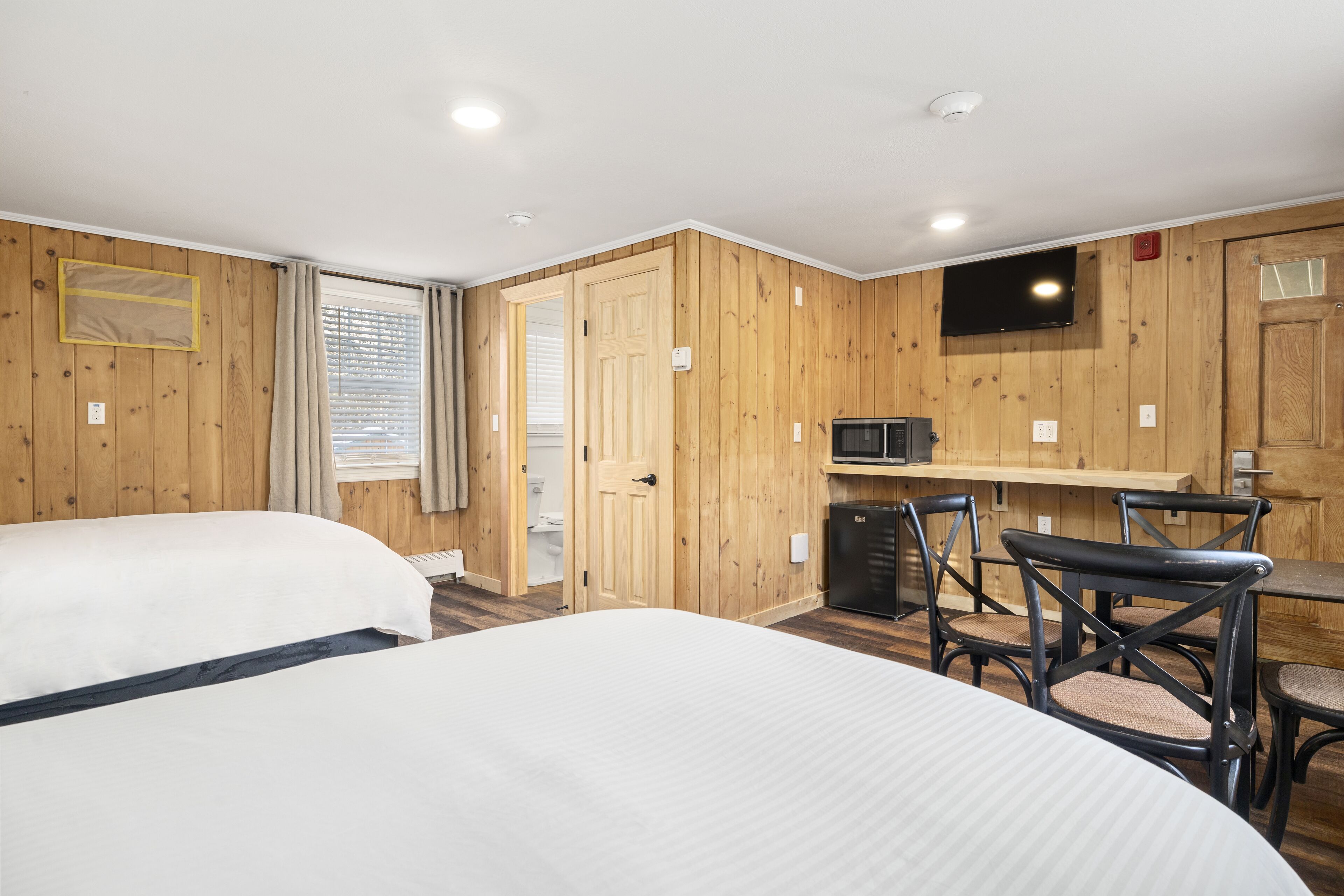 A hotel room with two beds, a wooden wall paneling, a television, a small kitchenette, and a dining area with chairs.