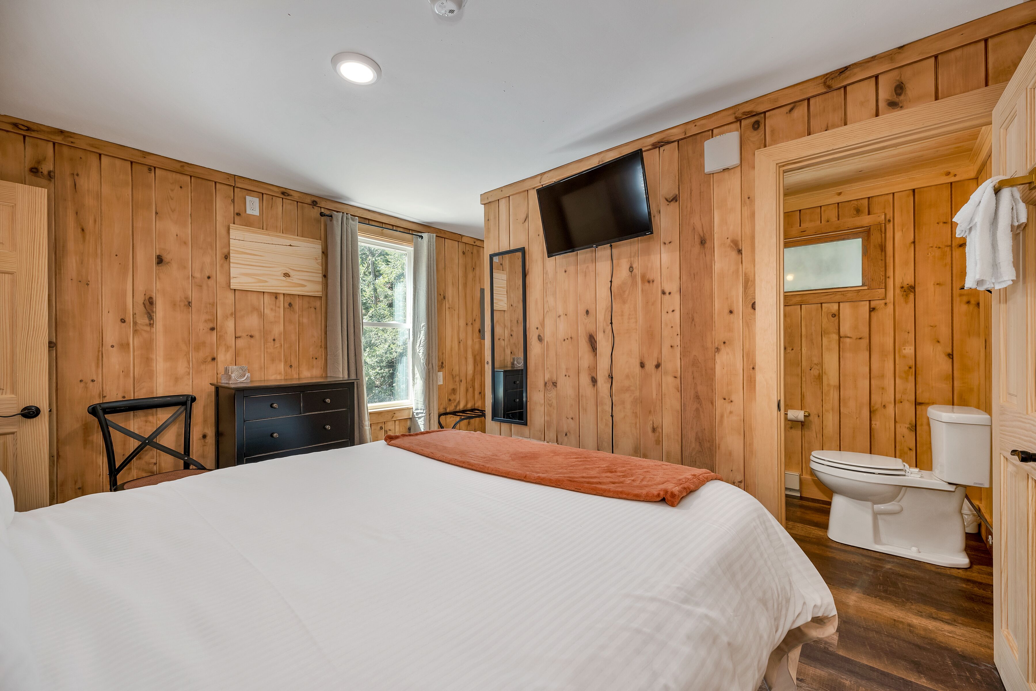 Una stanza con rivestimento in legno, un letto, una cassettiera, un televisore e un water.