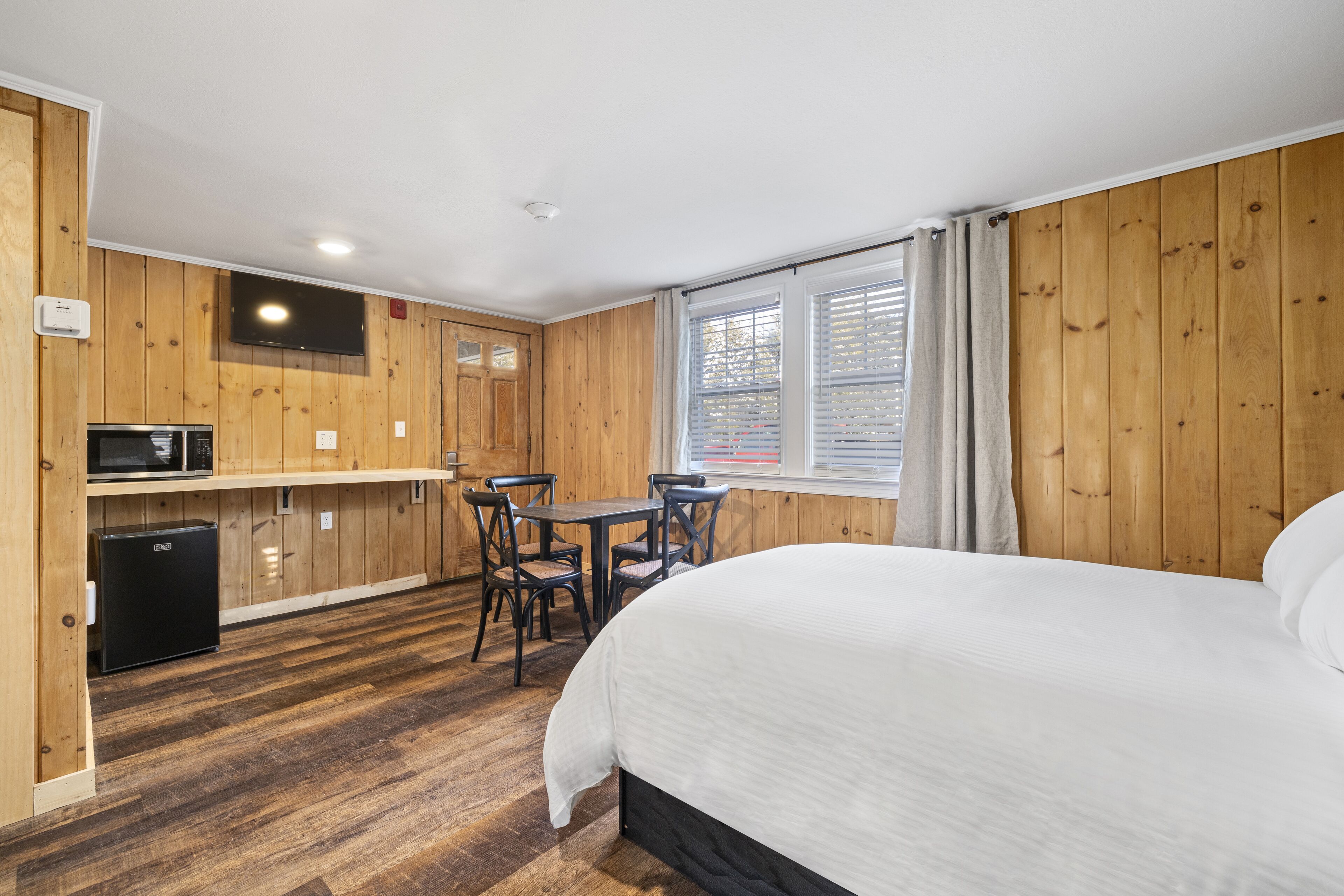 Een kamer met een bed, een kleine kitchenette, een eettafel met stoelen en een raam met gordijnen.