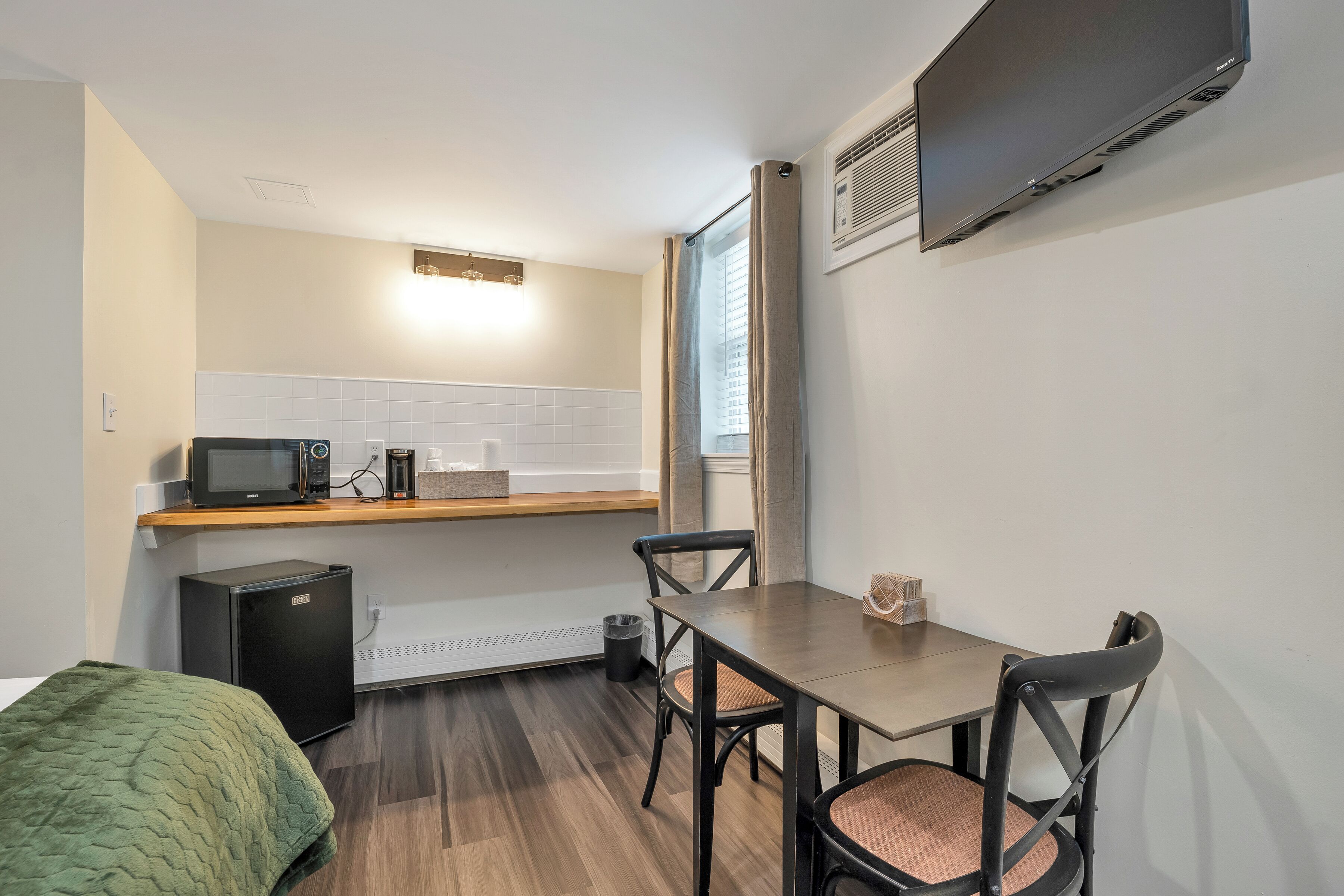 Una cocina compacta equipada con microondas, tostadora y refrigerador pequeño. Un comedor con sillas se encuentra cerca de una ventana con cortinas.
