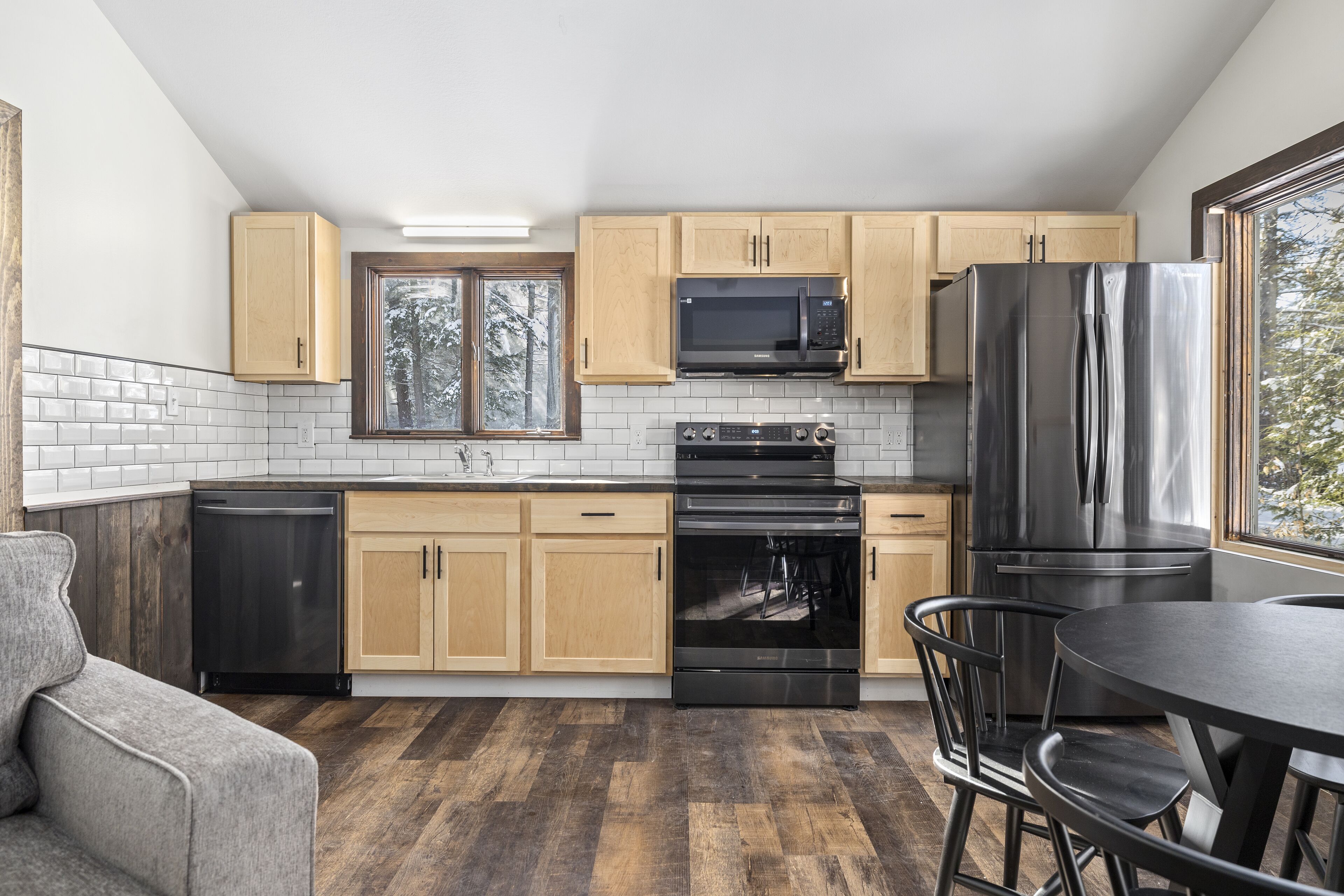 A modern kitchen with wooden cabinets, stainless steel appliances, a dining area with a table and chairs, and a view through the window.