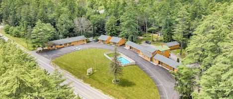 A lodge with multiple cabins, a swimming pool, and a parking area surrounded by dense forest and mountains.