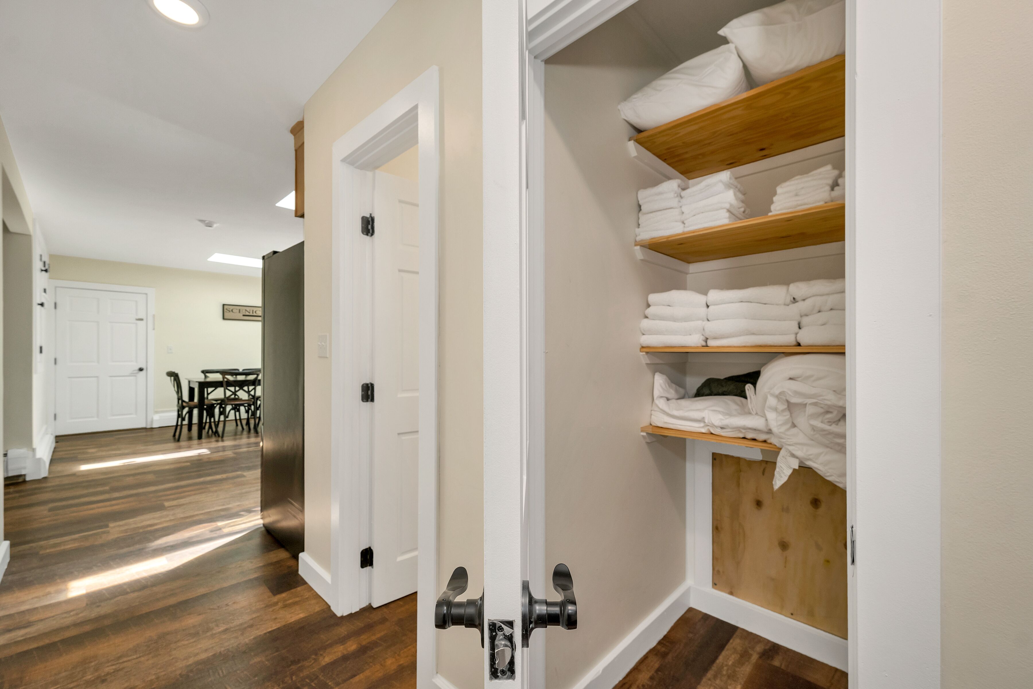 A hallway with wooden flooring leading to a room with a closet containing neatly folded towels.