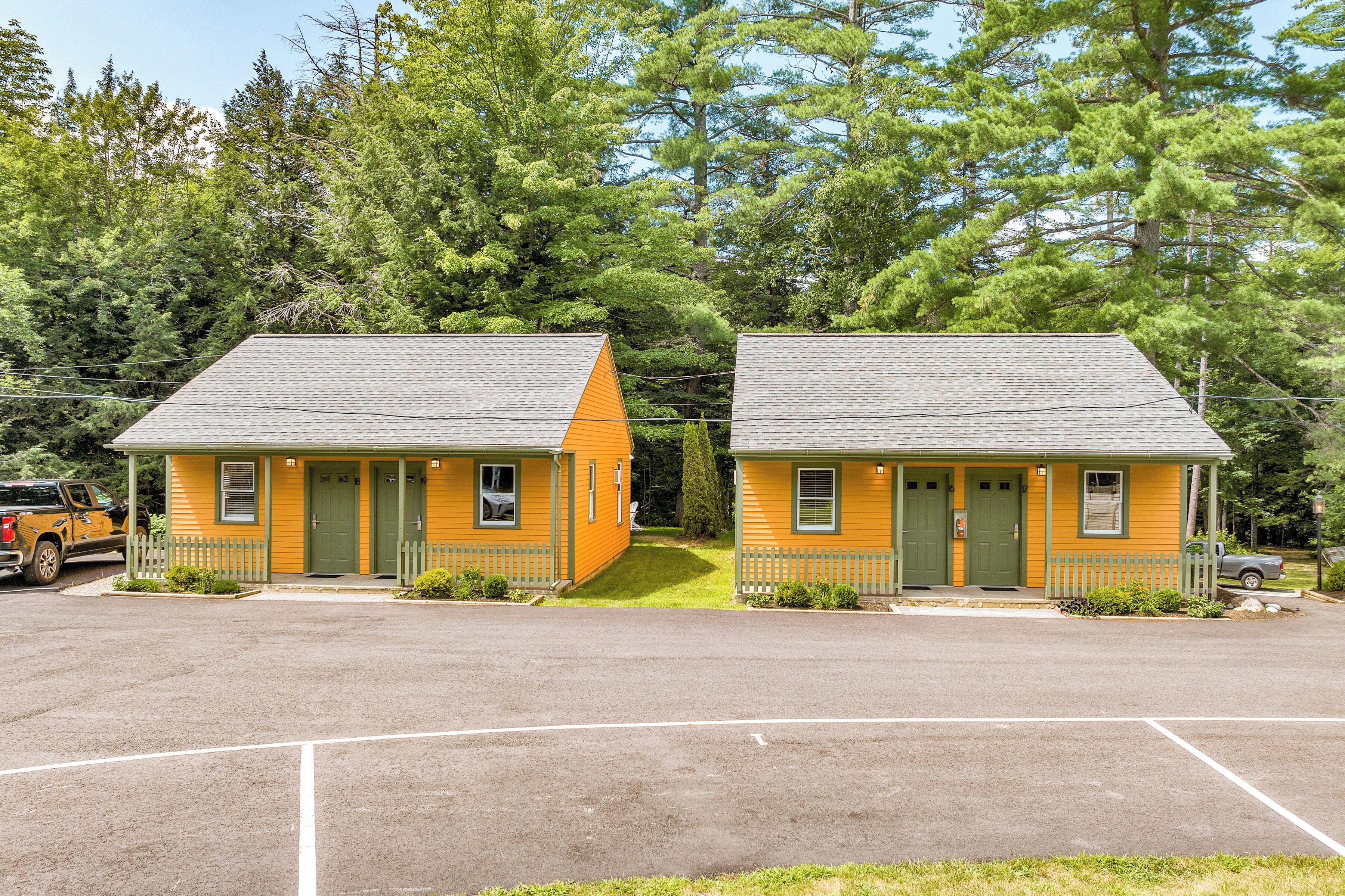 Two yellow cabins with green doors and windows, surrounded by a parking lot and trees.
