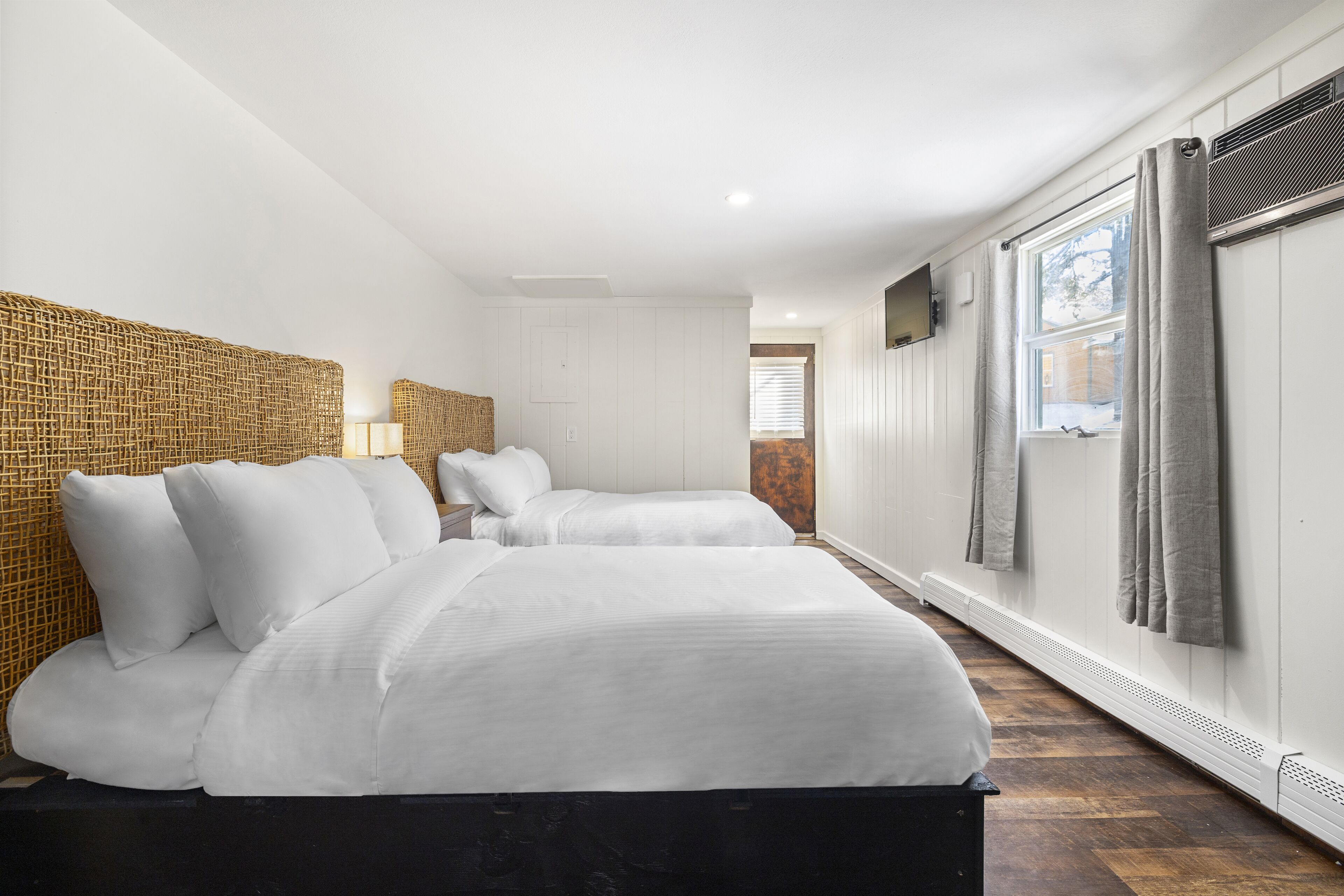 A hotel room with two beds, a woven headboard, a large window with curtains, and a radiator.