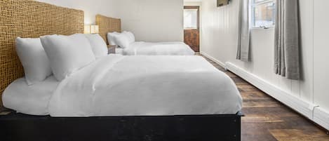 A hotel room with two beds, a woven headboard, a large window with curtains, and a radiator.